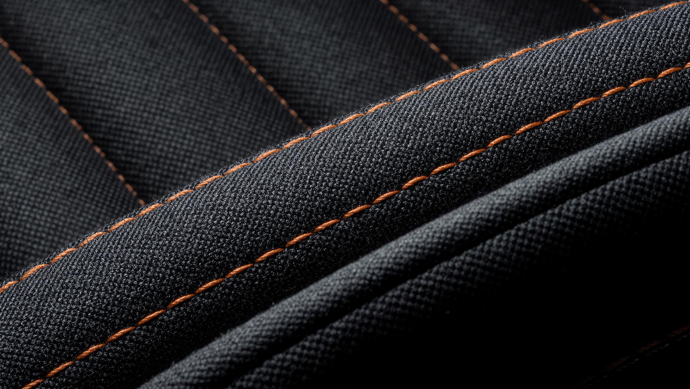 Macro view of a charcoal automotive seat fabric crossed by a double row of orange contrast topstitching, individual fibers visible in raking light.
