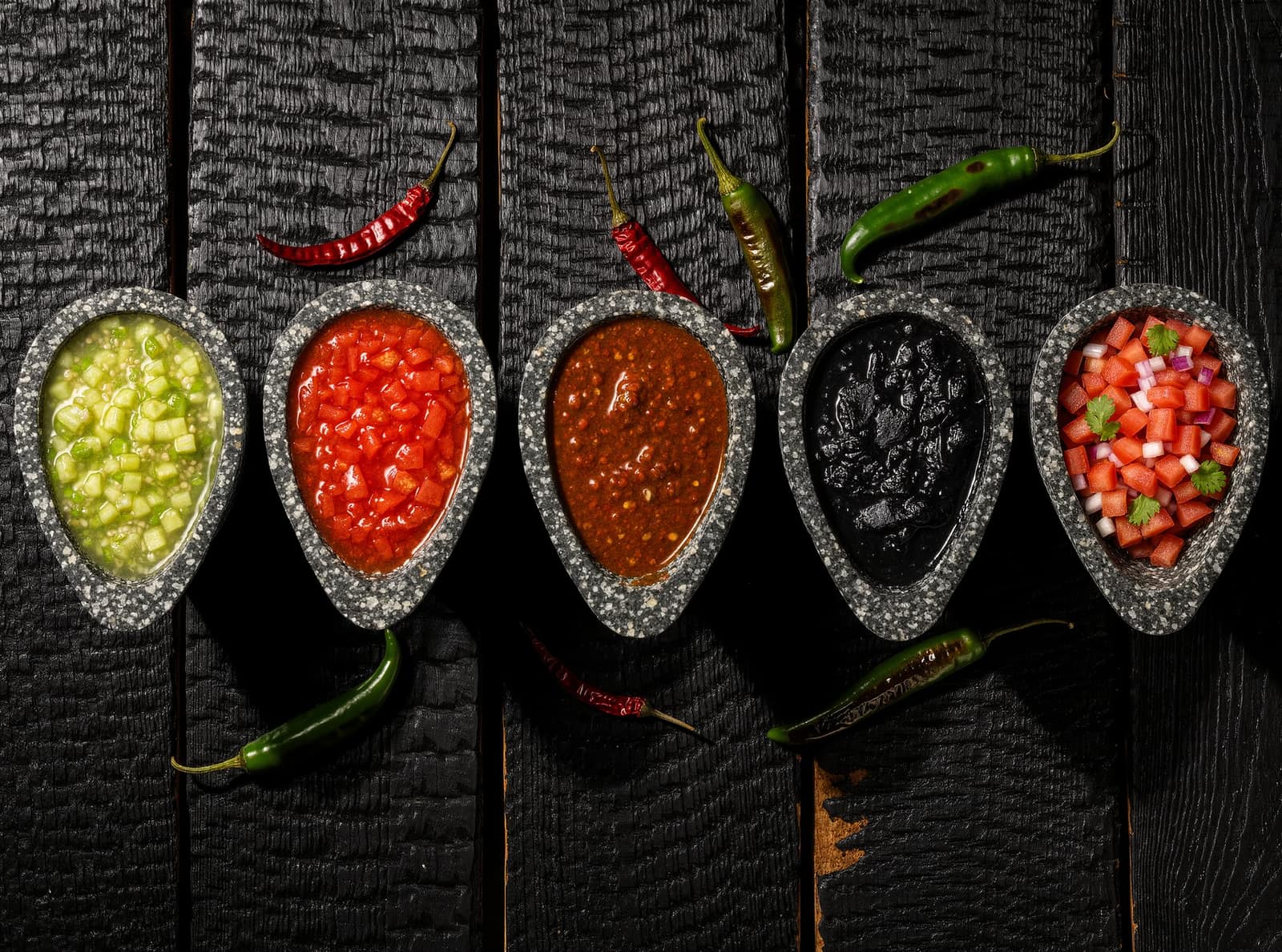Five stone molcajetes in a row on charred black wood, holding tomatillo green, bright red, deep brick, near-black charred salsa and chunky pico de gallo, with whole dried and fresh chiles scattered between them.