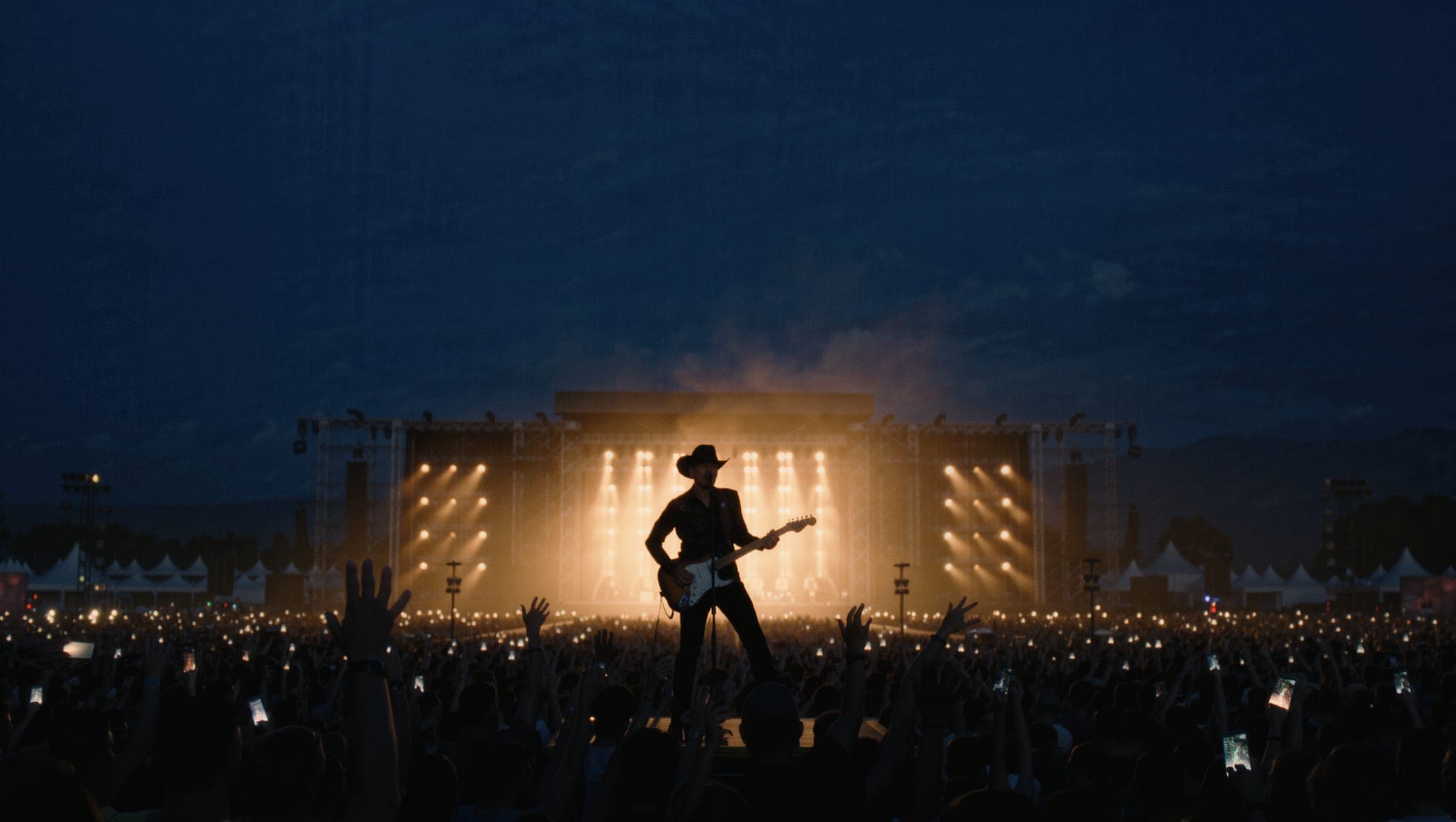 A festival stage at dusk seen from the crowd, the performer back-lit in amber and blue haze over thousands of raised hands.
