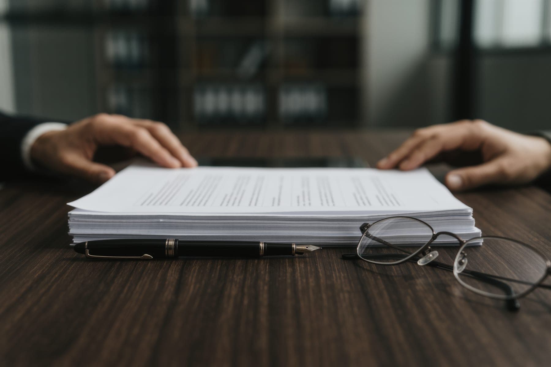 A dark walnut boardroom table with a stack of unmarked documents, a fountain pen, and two people's hands resting nearby.