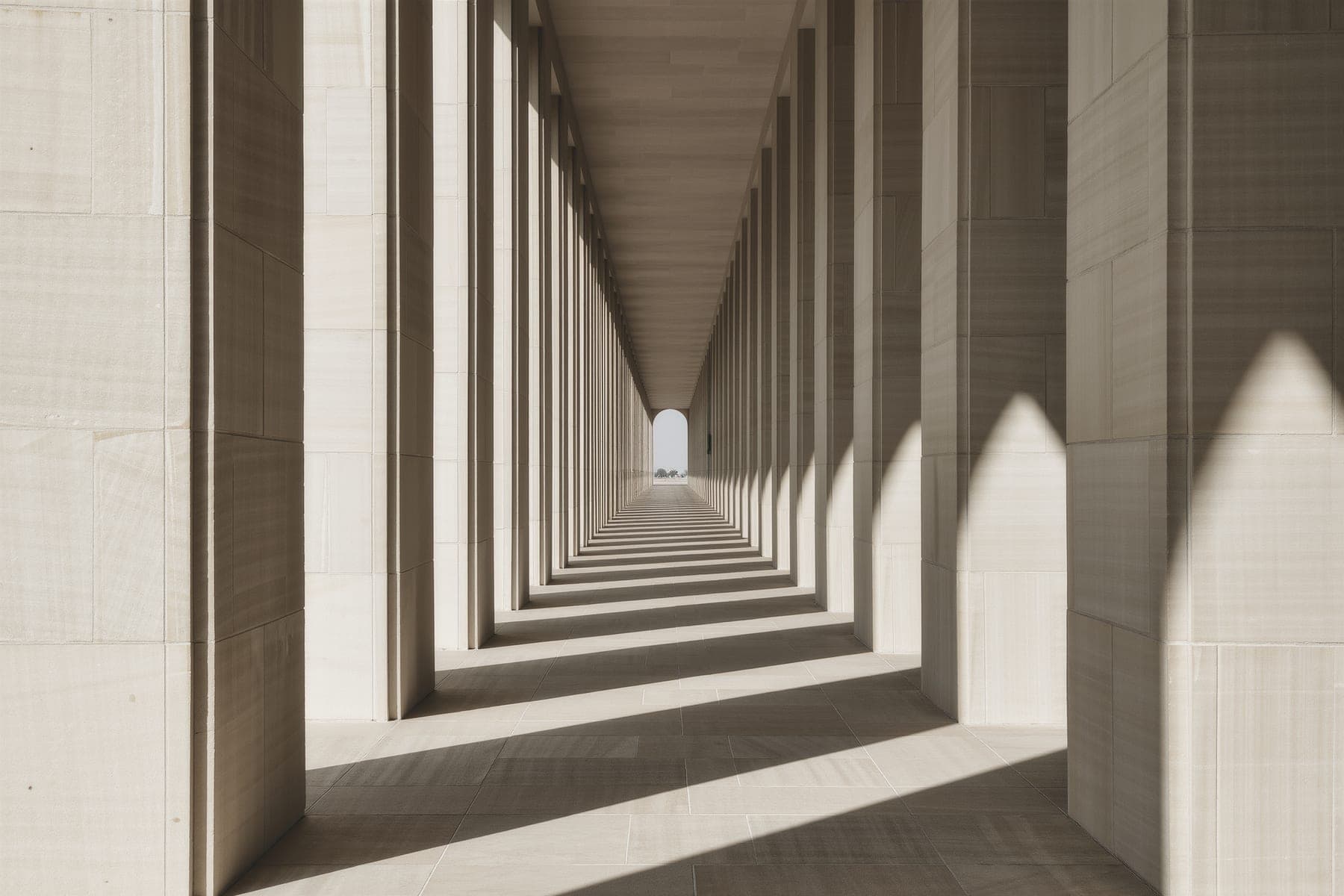 A colonnade of pale limestone piers in raking sunlight, hard geometric shadows across the floor.