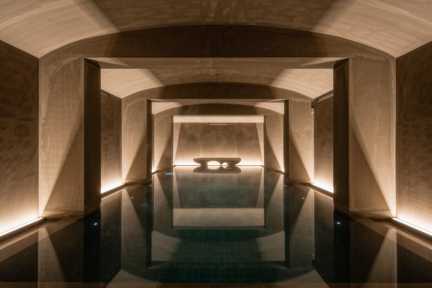A still indoor pool reflecting a vaulted concrete ceiling, with warm uplighting and faint steam.