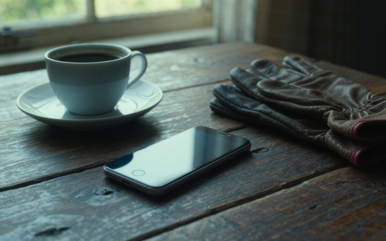 A smartphone lying face down on a weathered wooden table beside a ceramic coffee cup and leather work gloves in morning light