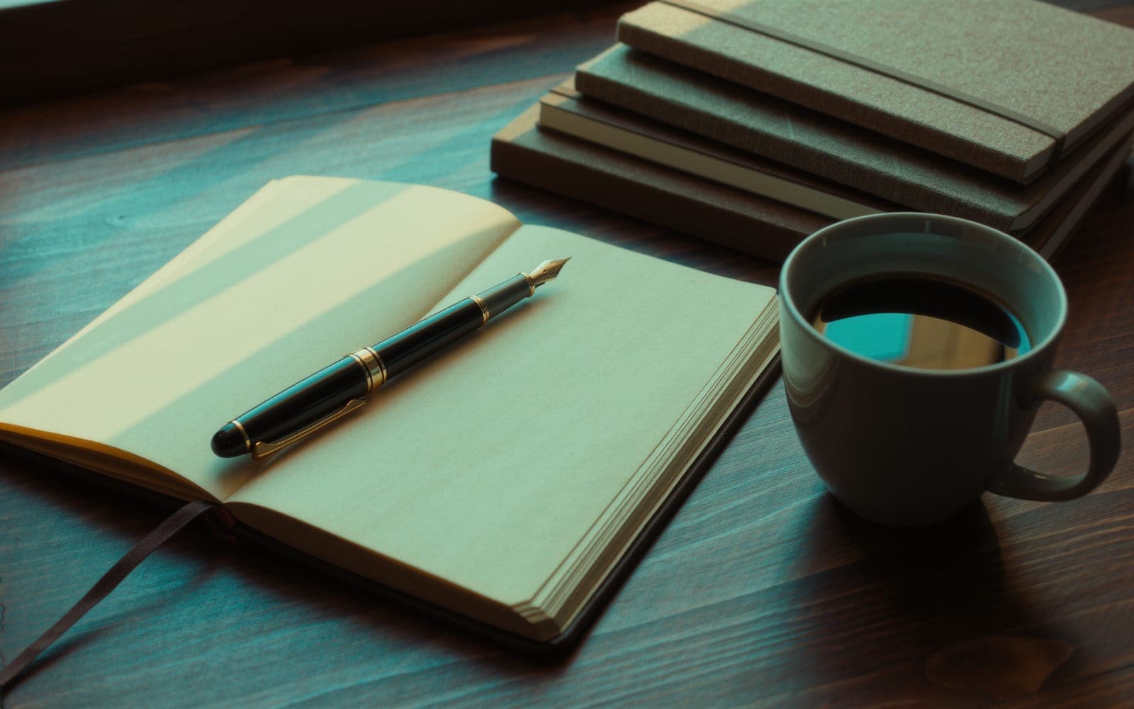 A fountain pen resting on an open cream notebook with blank pages beside a cup of black coffee and a stack of linen notebooks in morning light