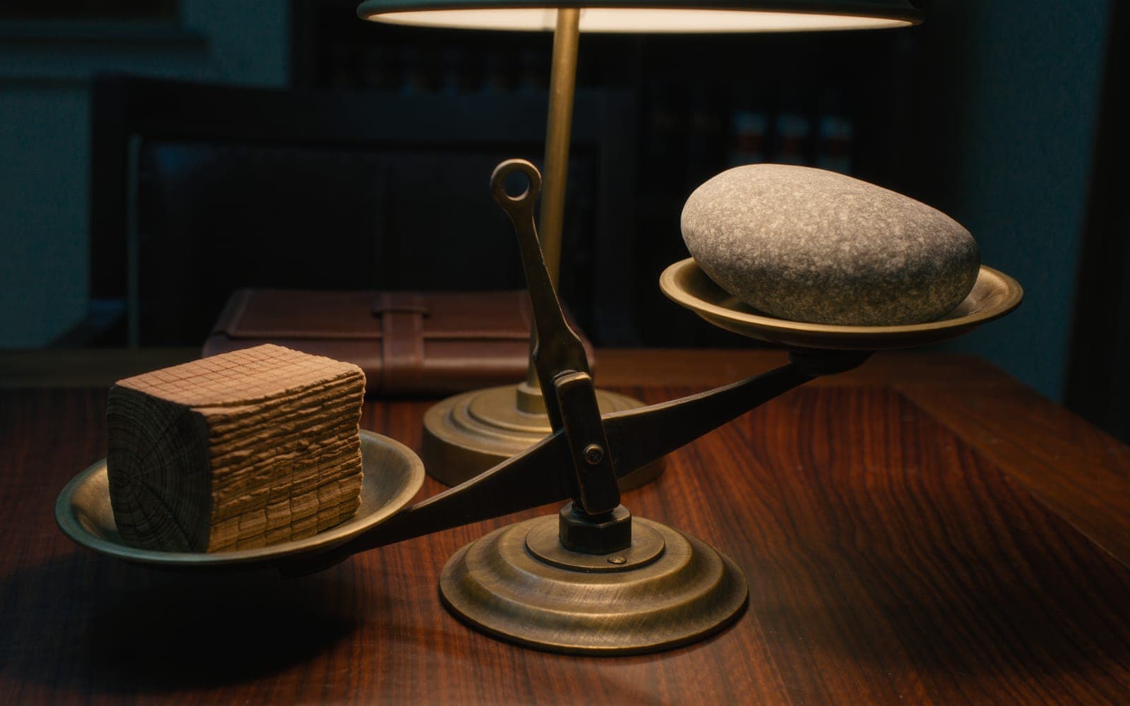 An antique brass balance scale on a walnut desk, a block of raw oak on one pan and a river stone on the other, under a warm lamp