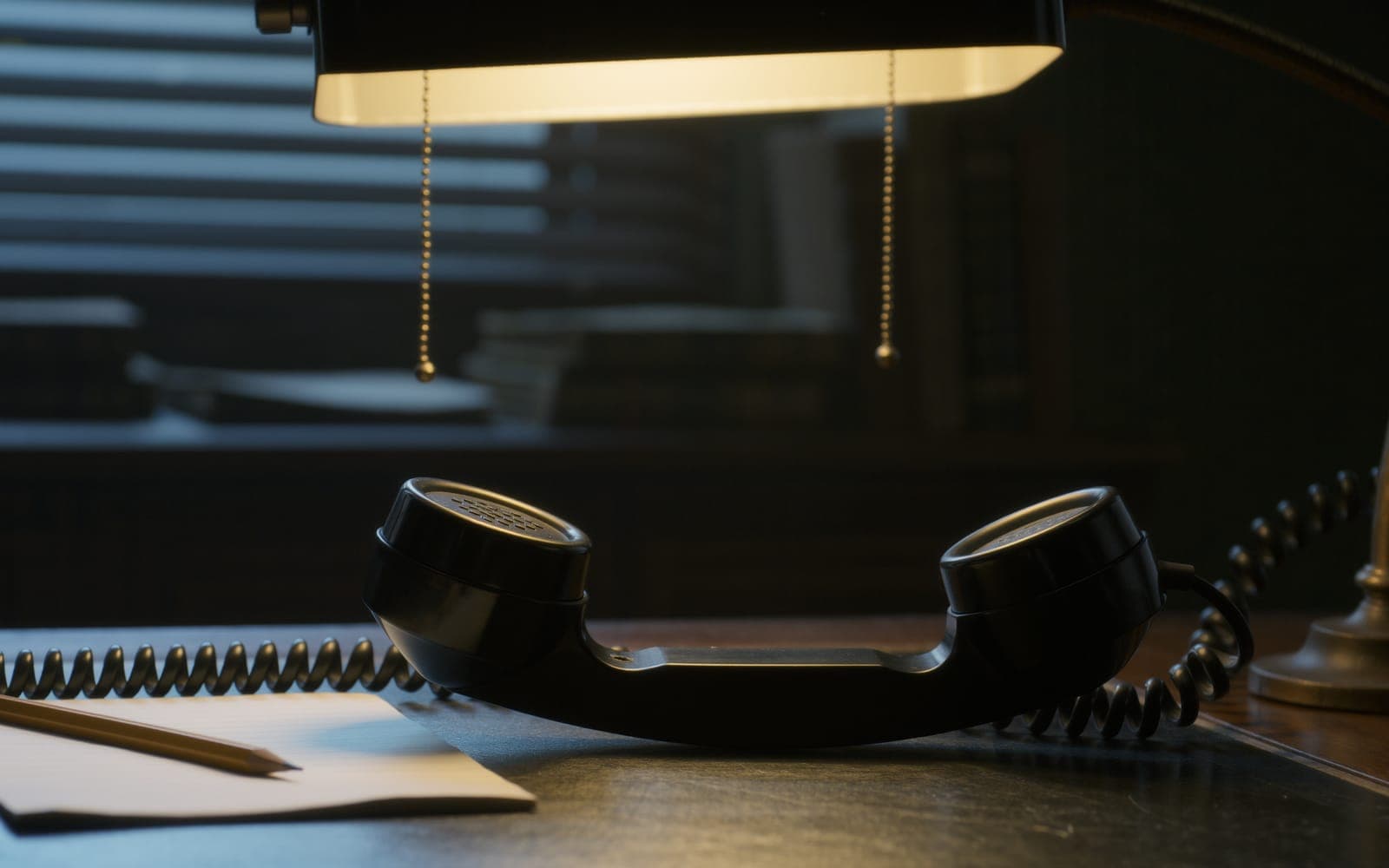 A black telephone handset lifted off its cradle on a dark wood desk beside a blank notepad and pencil under a warm lamp