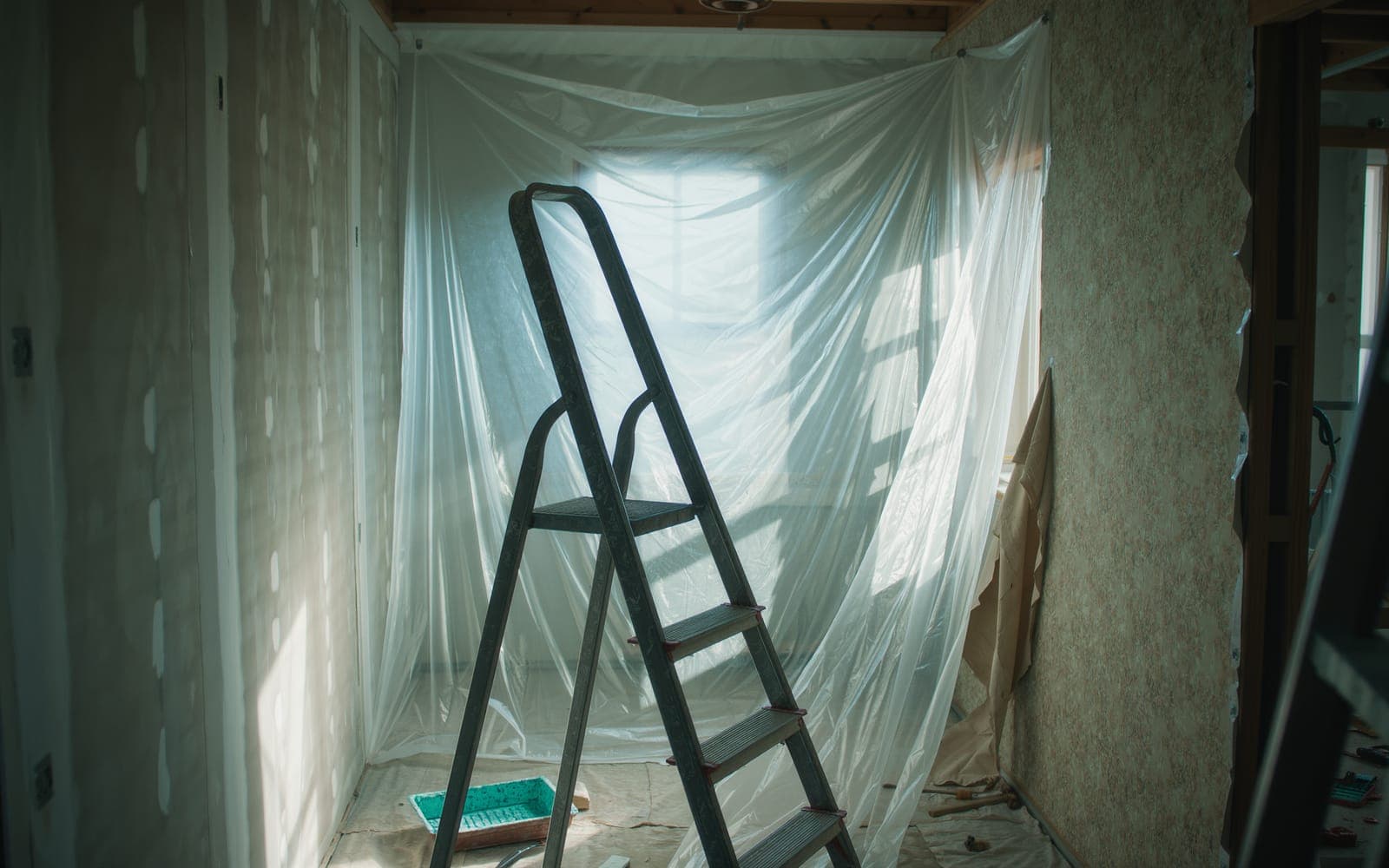 A room mid-renovation with fresh drywall beside peeling floral wallpaper, sunlight through plastic sheeting and a ladder