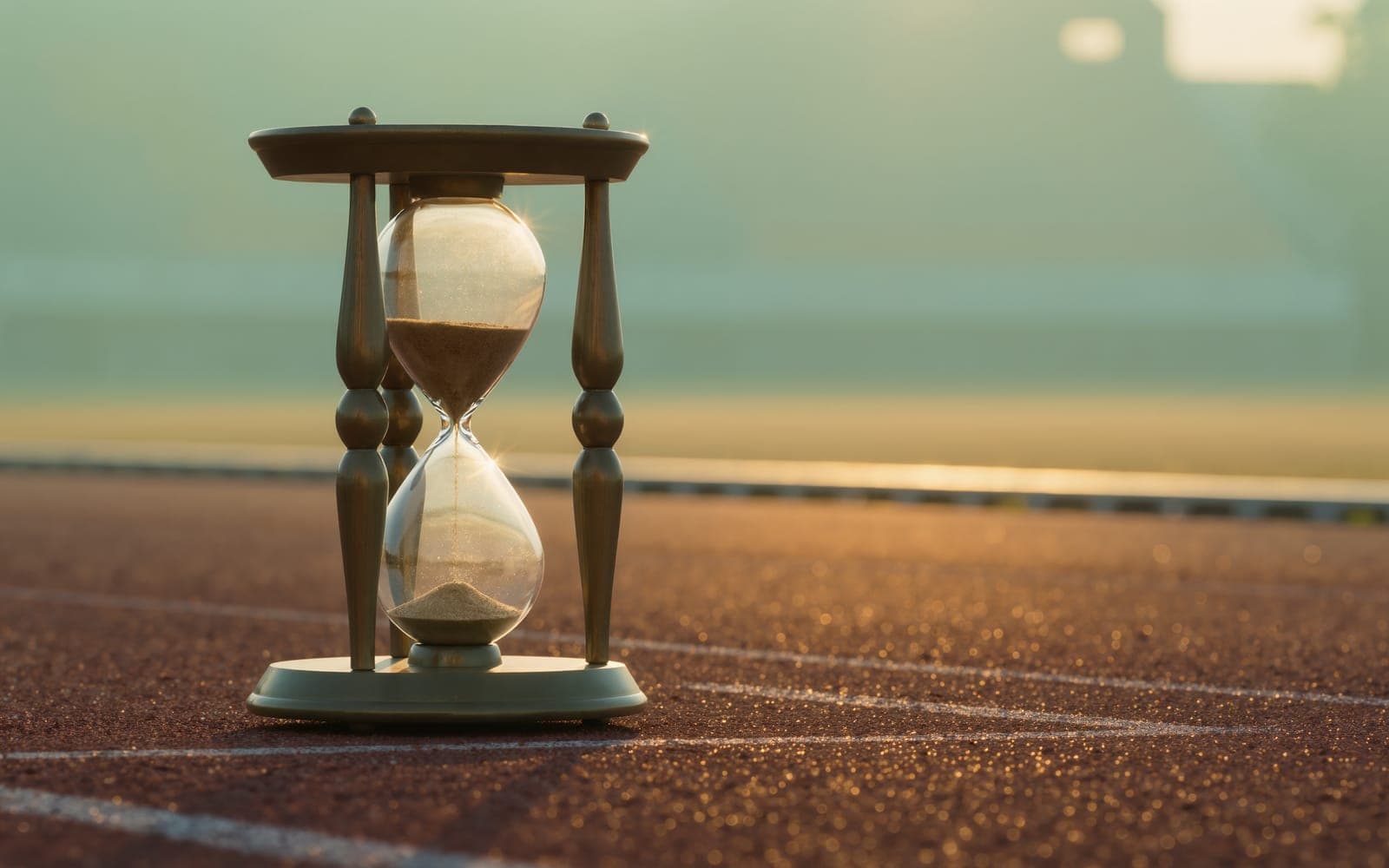 A brass-framed hourglass with amber sand mid-flow standing on a red running track starting line in morning mist
